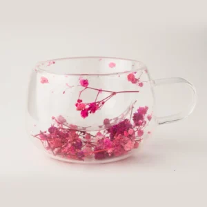 Caneca de vidro borossilicato transparente de parede dupla com flores secas naturais rosa e pink preservadas na base, criando efeito flutuante sobre fundo branco neutro